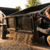 Contractor adjusting the spreader gate on a black dump trailer with gravel flowing out — dump trailer spreader gate calibration at PrimeLoad