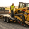 Contractor driving a CAT skid steer up the slide-in ramps onto a black 7x20 equipment trailer — how to load a skid steer at PrimeLoad