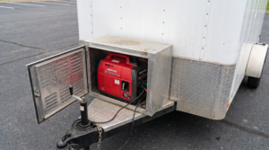 Sealed exterior generator box on the V-nose of a white enclosed cargo trailer with the door open showing an inverter generator inside — cargo trailer generator box at PrimeLoad